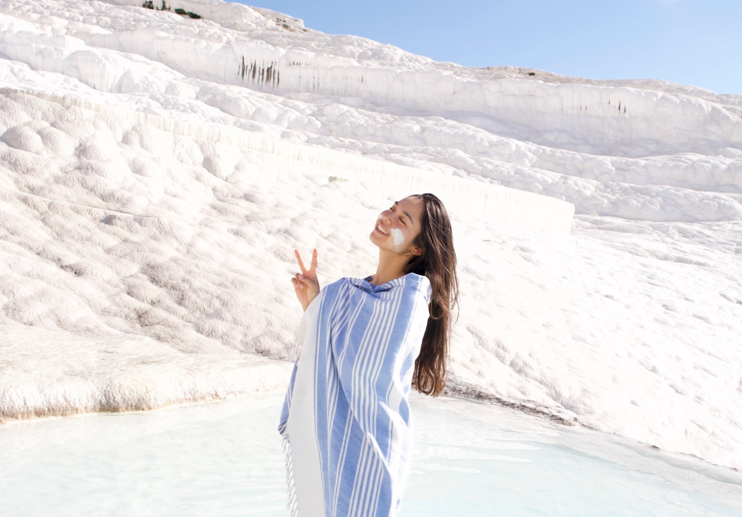 Selin the founder of SENDE smiling with the peace sign, standing in a clear thermal pool terrace on a sunny and bright day in Pamukkale, wearing a blue sea coloured SENDE peshtemal Turkish towel, and natural thermal clay as a skincare treatment.